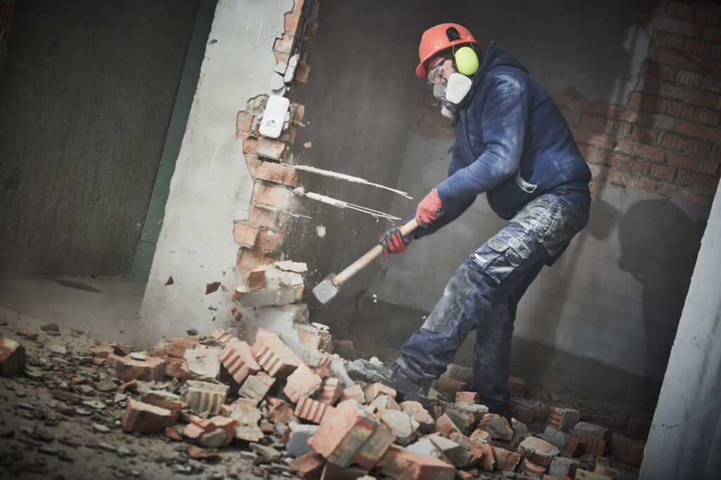 In Schutzkleidung zerstört ein Arbeiter geschickt mit einem Vorschlaghammer eine Ziegelwand im Innenbereich und geht dabei mit der Präzision eines Tatortreinigers an anspruchsvolle Aufräumarbeiten heran.